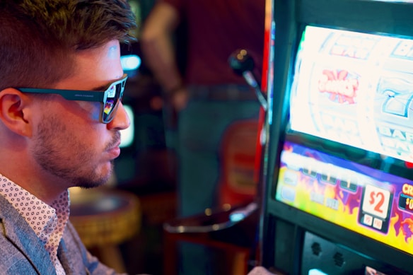 A man wearing sunglasses is intently focused on a slot machine, with colorful lights and graphics illuminating the screen. The scene is set indoors, likely in a casino or gaming area. His attire includes a patterned shirt and a blazer.