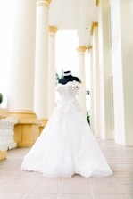 A mannequin displaying an elegant white wedding gown, featuring intricate lace details, stands in a corridor with tall cream-colored pillars. The setting is bright and airy, suggesting a grand architectural space.