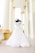 A mannequin displaying an elegant white wedding gown, featuring intricate lace details, stands in a corridor with tall cream-colored pillars. The setting is bright and airy, suggesting a grand architectural space.