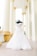 A mannequin displaying an elegant white wedding gown, featuring intricate lace details, stands in a corridor with tall cream-colored pillars. The setting is bright and airy, suggesting a grand architectural space.