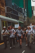 A group of people are marching in a city street, holding signs and banners with environmental messages. Buildings and a tram are visible in the background, suggesting an urban setting. Some individuals are using a megaphone to amplify their voices, indicating a protest or demonstration.