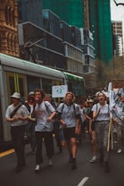 A group of people are marching in a city street, holding signs and banners with environmental messages. Buildings and a tram are visible in the background, suggesting an urban setting. Some individuals are using a megaphone to amplify their voices, indicating a protest or demonstration.