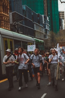 A group of people are marching in a city street, holding signs and banners with environmental messages. Buildings and a tram are visible in the background, suggesting an urban setting. Some individuals are using a megaphone to amplify their voices, indicating a protest or demonstration.