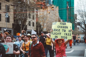 A group of people are participating in a climate change protest on a city street. Several individuals hold signs, including one prominently displayed that reads 'Cut your carbon? Pledge to be flight free in 2020.' The participants appear to be diverse in age and are dressed casually.