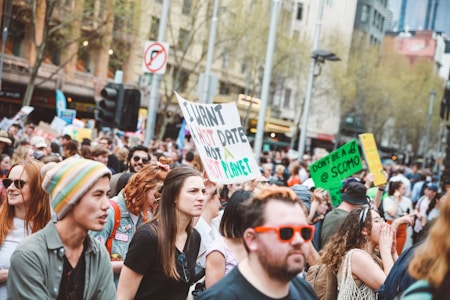A large group of people is gathered outdoors in a city setting, holding various signs protesting about climate change. The crowd includes diverse individuals, some wearing colorful hats and sunglasses. Trees and buildings are visible in the background, indicating an urban environment.
