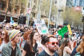 A large group of people is gathered outdoors in a city setting, holding various signs protesting about climate change. The crowd includes diverse individuals, some wearing colorful hats and sunglasses. Trees and buildings are visible in the background, indicating an urban environment.