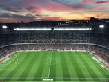 A large football stadium filled with spectators captures an ongoing match. The grass is impeccably maintained, and players in jerseys can be seen on the field. Above the stadium, a vibrant and colorful sunset paints the sky with hues of orange, pink, and purple, creating a stunning backdrop.