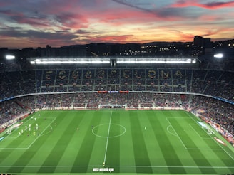 A large football stadium filled with spectators captures an ongoing match. The grass is impeccably maintained, and players in jerseys can be seen on the field. Above the stadium, a vibrant and colorful sunset paints the sky with hues of orange, pink, and purple, creating a stunning backdrop.