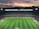 A large football stadium filled with spectators captures an ongoing match. The grass is impeccably maintained, and players in jerseys can be seen on the field. Above the stadium, a vibrant and colorful sunset paints the sky with hues of orange, pink, and purple, creating a stunning backdrop.