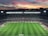 A large football stadium filled with spectators captures an ongoing match. The grass is impeccably maintained, and players in jerseys can be seen on the field. Above the stadium, a vibrant and colorful sunset paints the sky with hues of orange, pink, and purple, creating a stunning backdrop.