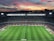 A large football stadium filled with spectators captures an ongoing match. The grass is impeccably maintained, and players in jerseys can be seen on the field. Above the stadium, a vibrant and colorful sunset paints the sky with hues of orange, pink, and purple, creating a stunning backdrop.