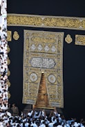 A large group of individuals dressed in white gather around the ornate, gold-embroidered door of the Kaaba. The black fabric covering the Kaaba is adorned with intricate Arabic calligraphy in gold.