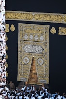 A large group of individuals dressed in white gather around the ornate, gold-embroidered door of the Kaaba. The black fabric covering the Kaaba is adorned with intricate Arabic calligraphy in gold.