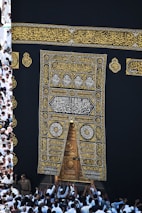 A large group of individuals dressed in white gather around the ornate, gold-embroidered door of the Kaaba. The black fabric covering the Kaaba is adorned with intricate Arabic calligraphy in gold.