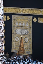 A large group of individuals dressed in white gather around the ornate, gold-embroidered door of the Kaaba. The black fabric covering the Kaaba is adorned with intricate Arabic calligraphy in gold.