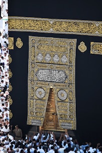 A large group of individuals dressed in white gather around the ornate, gold-embroidered door of the Kaaba. The black fabric covering the Kaaba is adorned with intricate Arabic calligraphy in gold.