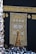 A large group of individuals dressed in white gather around the ornate, gold-embroidered door of the Kaaba. The black fabric covering the Kaaba is adorned with intricate Arabic calligraphy in gold.