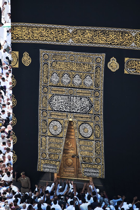 A large group of individuals dressed in white gather around the ornate, gold-embroidered door of the Kaaba. The black fabric covering the Kaaba is adorned with intricate Arabic calligraphy in gold.
