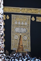 A large group of individuals dressed in white gather around the ornate, gold-embroidered door of the Kaaba. The black fabric covering the Kaaba is adorned with intricate Arabic calligraphy in gold.
