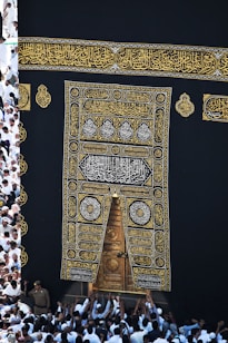 A large group of individuals dressed in white gather around the ornate, gold-embroidered door of the Kaaba. The black fabric covering the Kaaba is adorned with intricate Arabic calligraphy in gold.