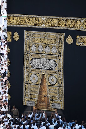 A large group of individuals dressed in white gather around the ornate, gold-embroidered door of the Kaaba. The black fabric covering the Kaaba is adorned with intricate Arabic calligraphy in gold.