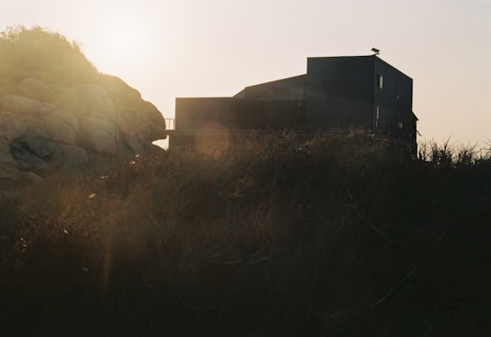 A modern, dark-colored house appears against the backdrop of a bright, hazy sky. The scene is framed by foliage and rocky terrain, with sunlight casting a soft glow across the image.