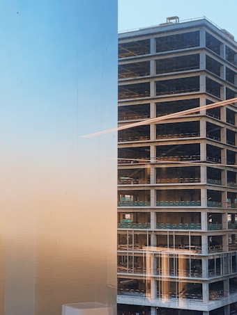 A high-rise building under construction is captured during the early evening. The structure's concrete framework is visible, with scaffolding and open floors. The left side of the image reflects the sunset, presenting a gradient of blue and orange colors.