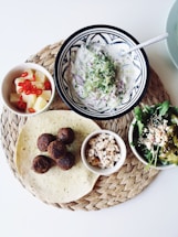 A woven placemat holds a plate of falafel balls, a tortilla, a bowl of flavorful salad with greens and peppers, a small dish of crumbled cheese, a bowl of creamy yogurt dip, and a cup of pineapple cubes with sliced red chilies. The display is neatly arranged against a white background, capturing a Mediterranean or Middle Eastern culinary theme.