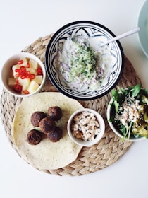 A woven placemat holds a plate of falafel balls, a tortilla, a bowl of flavorful salad with greens and peppers, a small dish of crumbled cheese, a bowl of creamy yogurt dip, and a cup of pineapple cubes with sliced red chilies. The display is neatly arranged against a white background, capturing a Mediterranean or Middle Eastern culinary theme.