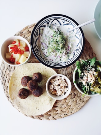 A woven placemat holds a plate of falafel balls, a tortilla, a bowl of flavorful salad with greens and peppers, a small dish of crumbled cheese, a bowl of creamy yogurt dip, and a cup of pineapple cubes with sliced red chilies. The display is neatly arranged against a white background, capturing a Mediterranean or Middle Eastern culinary theme.