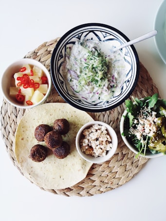 A woven placemat holds a plate of falafel balls, a tortilla, a bowl of flavorful salad with greens and peppers, a small dish of crumbled cheese, a bowl of creamy yogurt dip, and a cup of pineapple cubes with sliced red chilies. The display is neatly arranged against a white background, capturing a Mediterranean or Middle Eastern culinary theme.