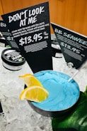 A display of skincare products set on ice, featuring a bowl of bright blue cleansing mask with a metal spoon. Two slices of lemon garnish the edge of the bowl. Behind it, black signs with white text provide information about the products, highlighting a product named 'Don't Look at Me' priced at $13.95 for 60g.