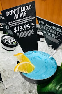 A display of skincare products set on ice, featuring a bowl of bright blue cleansing mask with a metal spoon. Two slices of lemon garnish the edge of the bowl. Behind it, black signs with white text provide information about the products, highlighting a product named 'Don't Look at Me' priced at $13.95 for 60g.