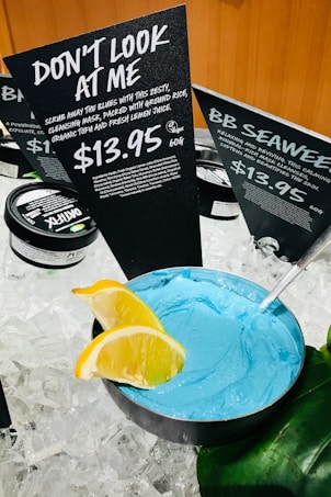 A display of skincare products set on ice, featuring a bowl of bright blue cleansing mask with a metal spoon. Two slices of lemon garnish the edge of the bowl. Behind it, black signs with white text provide information about the products, highlighting a product named 'Don't Look at Me' priced at $13.95 for 60g.