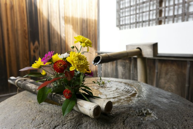 A rustic setting featuring a traditional bamboo fountain with water gently pouring into a stone basin. A vibrant bouquet of flowers with shades of yellow, red, white, and purple is placed on the bamboo structure. The surroundings have a warm, wooden texture with subtle lighting.