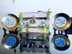 Two decorative plates with intricate Arabic calligraphy are placed on black stands, flanking a wooden crate adorned with artificial green vines and yellow flowers. The plates are mirrored on a glossy surface, enhancing their ornate golden designs.