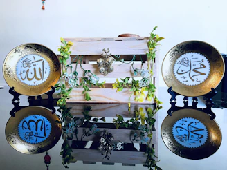 Two decorative plates with intricate Arabic calligraphy are placed on black stands, flanking a wooden crate adorned with artificial green vines and yellow flowers. The plates are mirrored on a glossy surface, enhancing their ornate golden designs.