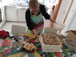 A person wearing a colorful apron is decorating a large rectangular cake with a bowl of cream in a kitchen setting. The cake is placed on a vibrant, patterned tablecloth, and there are various kitchen items scattered around, including a glass, a dish with pastry pieces, a red cloth, and a smartphone.
