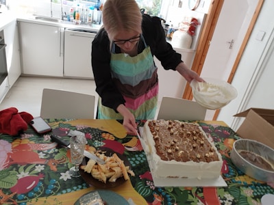 A person wearing a colorful apron is decorating a large rectangular cake with a bowl of cream in a kitchen setting. The cake is placed on a vibrant, patterned tablecloth, and there are various kitchen items scattered around, including a glass, a dish with pastry pieces, a red cloth, and a smartphone.