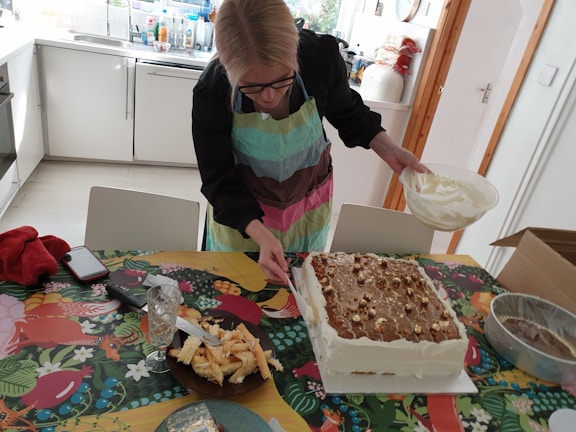 A person wearing a colorful apron is decorating a large rectangular cake with a bowl of cream in a kitchen setting. The cake is placed on a vibrant, patterned tablecloth, and there are various kitchen items scattered around, including a glass, a dish with pastry pieces, a red cloth, and a smartphone.