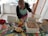 A person wearing a colorful apron is decorating a large rectangular cake with a bowl of cream in a kitchen setting. The cake is placed on a vibrant, patterned tablecloth, and there are various kitchen items scattered around, including a glass, a dish with pastry pieces, a red cloth, and a smartphone.