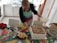 A person wearing a colorful apron is decorating a large rectangular cake with a bowl of cream in a kitchen setting. The cake is placed on a vibrant, patterned tablecloth, and there are various kitchen items scattered around, including a glass, a dish with pastry pieces, a red cloth, and a smartphone.