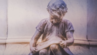 A stone sculpture of a child sitting and reading a book, with a soft and dreamy lighting creating a contemplative and serene atmosphere.