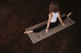 A person is practicing yoga on a mat in a dimly lit room with wooden floors. The person is wearing a white sleeveless top and black pants, sitting in a split position with hands on the mat, facing away. The mat has artistic designs.