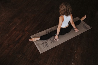 A person is practicing yoga on a mat in a dimly lit room with wooden floors. The person is wearing a white sleeveless top and black pants, sitting in a split position with hands on the mat, facing away. The mat has artistic designs.