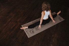 A person is practicing yoga on a mat in a dimly lit room with wooden floors. The person is wearing a white sleeveless top and black pants, sitting in a split position with hands on the mat, facing away. The mat has artistic designs.