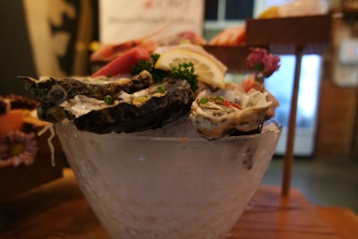 A bowl of fresh oysters garnished with slices of lemon, green herbs, and chili peppers, placed on a bed of ice. In the background, there are blurred images of other food items displayed, and the setting appears to be indoors with soft lighting.