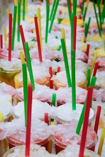 A collection of colorful drinks topped with crushed ice, each cup featuring a combination of red, green, and yellow straws. The assortment of colors suggests a variety of flavors, evoking a sense of freshness and summer.