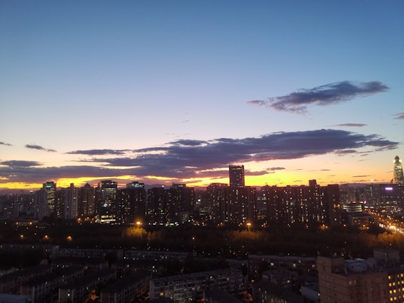 A city skyline is silhouetted against a vibrant sunset, with high-rise buildings creating a dark outline. The sky transitions from a bright orange and pink near the horizon to a deep blue above, scattered with a few dark clouds. Streetlights and building lights are visible as the city begins to light up for the evening.