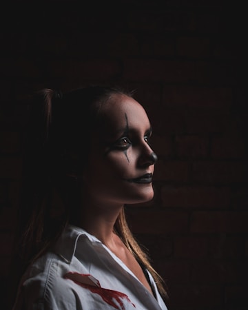A person with dark, theatrical makeup is captured in profile against a dimly lit background. The makeup features exaggerated black lines and a stark white base, giving a dramatic, intense look. The individual is wearing a white garment, possibly a shirt, with noticeable red stains, enhancing the dramatic and possibly eerie atmosphere.